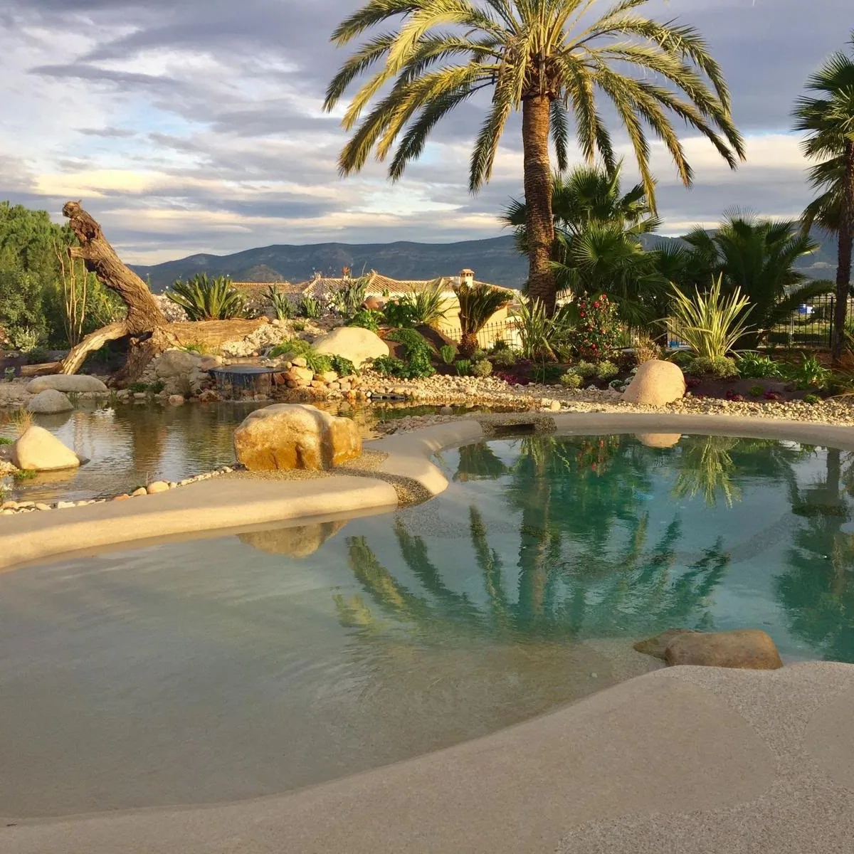 Piscina de arena tipo laguna al atardecer, con playa de entrada, rocas y palmeras